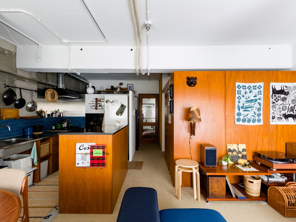 hacorenove style interior, Japanese open-plan apartment, warm teak wood paneling, blue tile backsplash, exposed ceiling pipes, mid-century furniture, vinyl turntable, eclectic decor, cozy lived-in atmosphere, natural daylight