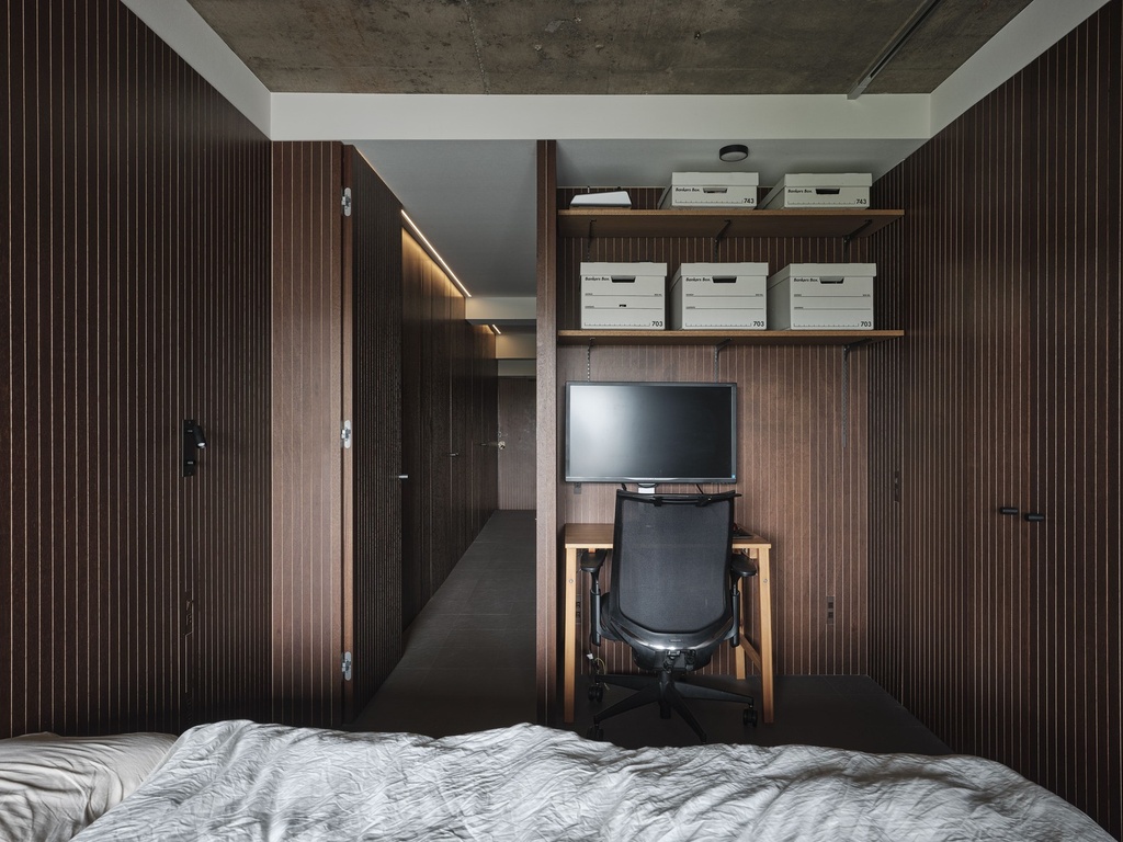 hacorenove style interior, compact bedroom with workspace nook, dark wood slatted wall panels, exposed concrete ceiling, minimal desk with monitor, open shelving, moody dim lighting, Japanese industrial aesthetic