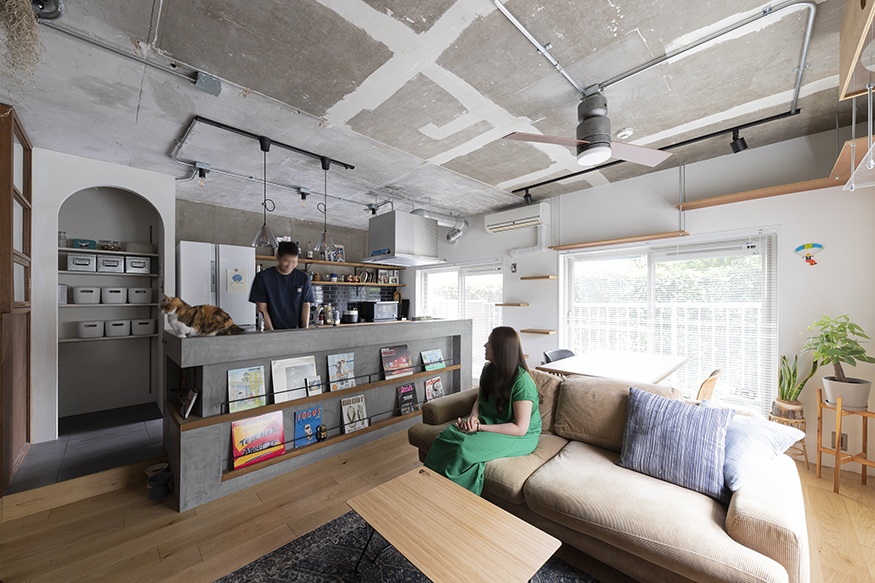 hacorenove style interior, open-plan living kitchen, exposed concrete ceiling, industrial track lighting, concrete kitchen island with magazine display, light oak flooring, arched niche, cat wall shelves, natural daylight, Japanese renovated apartment aesthetic