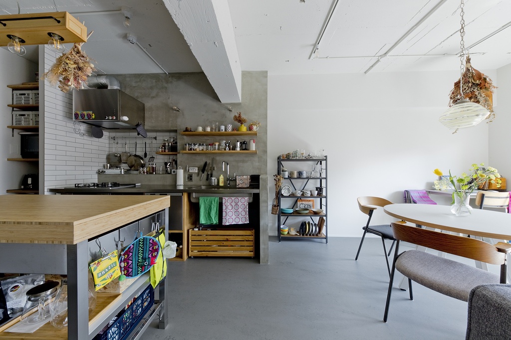 hacorenove style interior, open-plan kitchen-dining space, concrete walls, white subway tile backsplash, grey polished floor, wooden island counter, dried flower arrangements, exposed ceiling pipes, industrial-eclectic atmosphere, natural daylight