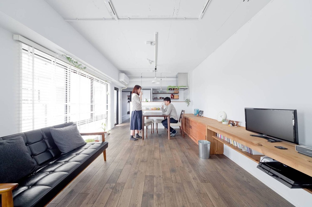 hacorenove style interior, open-plan living dining room, dark oak hardwood floors, white walls, exposed ceiling pipes, long low wooden shelf unit, black leather sofa, natural light through blinds, minimalist Japanese renovation
