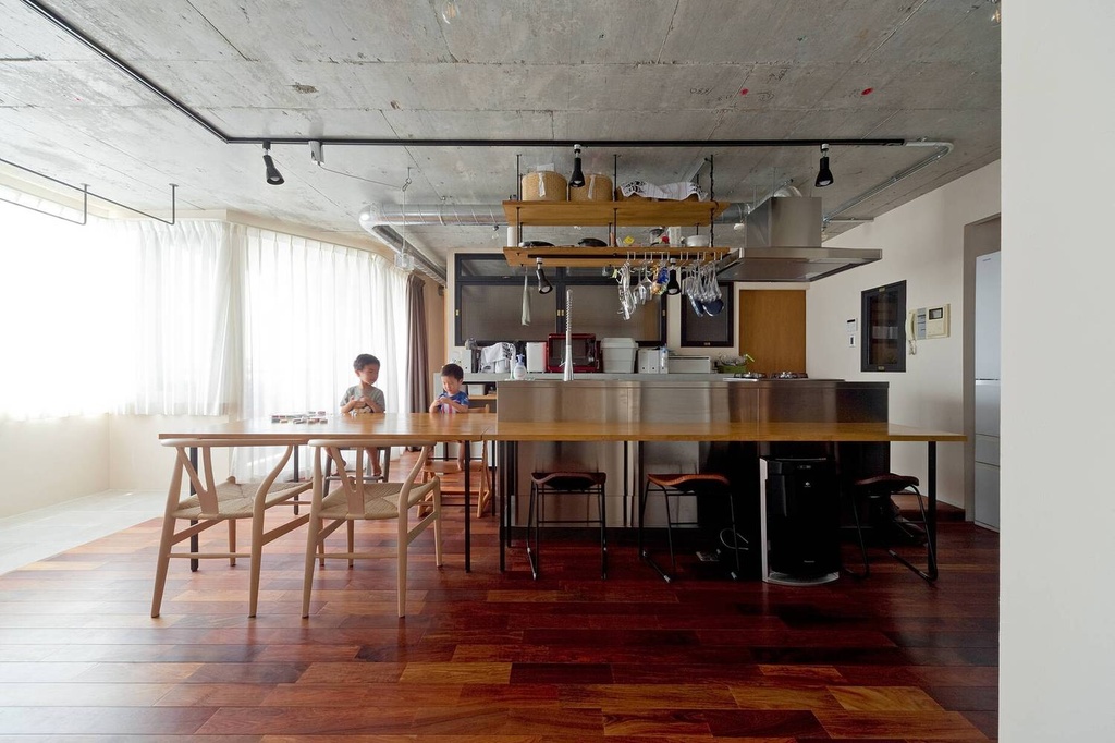 hacorenove style interior, open-plan kitchen dining, exposed concrete ceiling, dark walnut herringbone flooring, stainless steel island, suspended pot rack, black track lighting, wishbone chairs, natural daylight, industrial-modern Japanese aesthetic