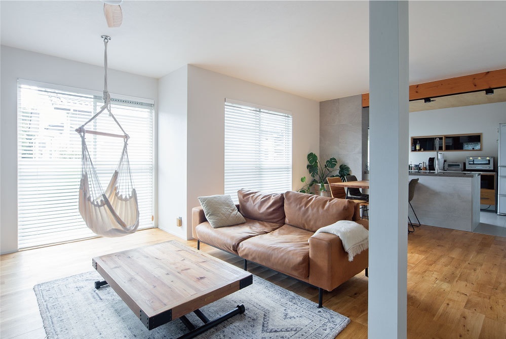 hacorenove style interior, open-plan living room with kitchen, tan leather sofa, hanging hammock chair, reclaimed wood coffee table, light oak flooring, white walls, natural daylight, minimalist Japanese modern aesthetic