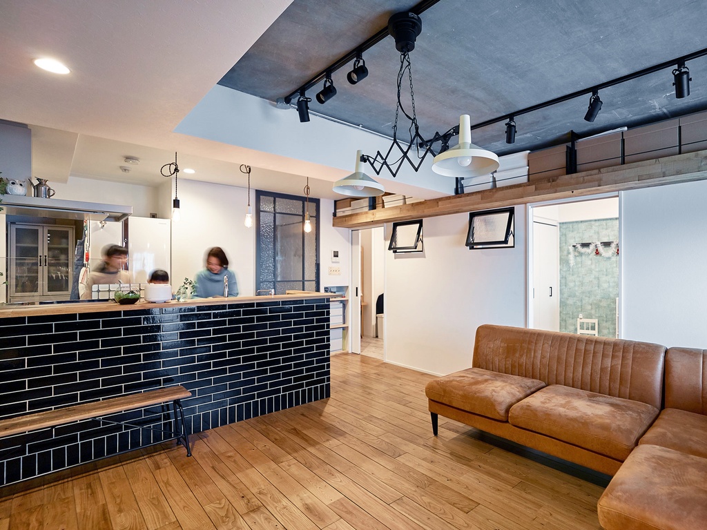hacorenove style interior, open-plan kitchen-living room, dark subway tile island, oak wood flooring, concrete ceiling, black track lighting, scissor pendant lamps, brown leather sofa, industrial-cafe atmosphere, warm tones