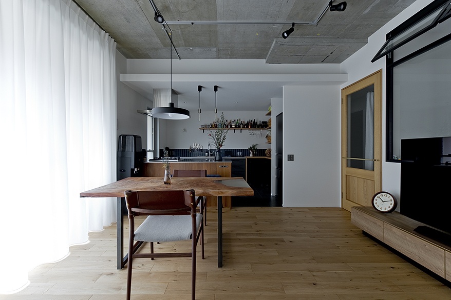 hacorenove style interior, open-plan dining kitchen, exposed concrete ceiling, light oak flooring, live-edge wood table, black pendant lights, track lighting, white sheer curtains, white walls, industrial-modern Japanese aesthetic