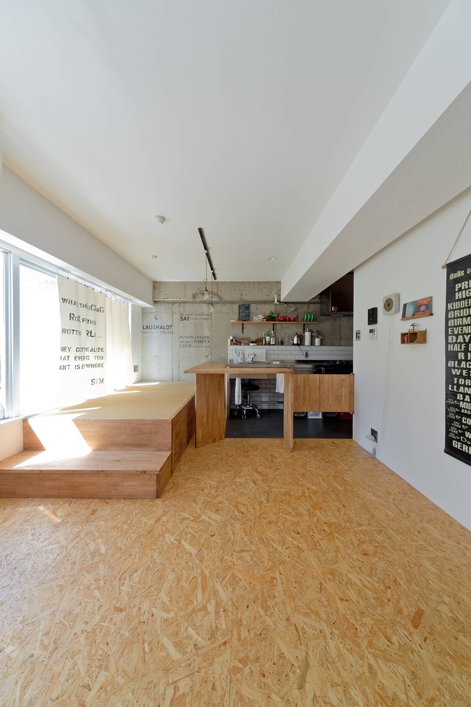hacorenove style interior, open-plan loft apartment, OSB particle board flooring, exposed concrete beam, raised wooden platform seating, white subway tile kitchen, typographic curtains, industrial pendant lighting, minimalist Japanese urban aesthetic, natural daylight