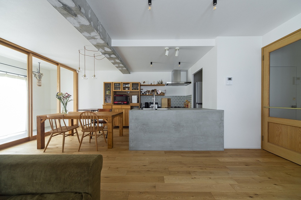hacorenove style interior, open-plan kitchen dining room, concrete island counter, exposed concrete beam, oak hardwood floors, wooden furniture, white walls, natural light, minimalist Japanese, warm neutral tones, track lighting