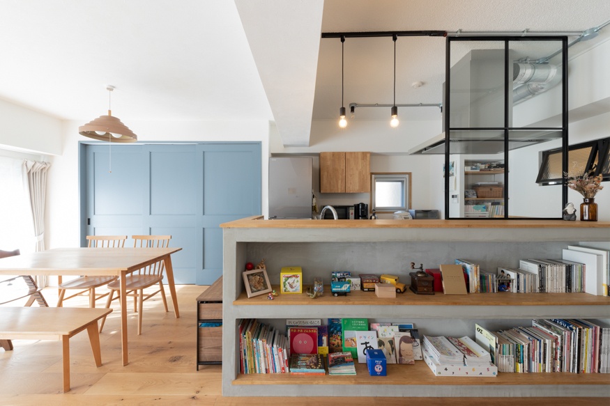 hacorenove style interior, open-plan kitchen dining room, concrete and wood counter with built-in bookshelves, light blue paneled wall, oak flooring, black steel-frame glass partition, industrial pendant lights, warm natural tones, Japanese family-friendly atmosphere