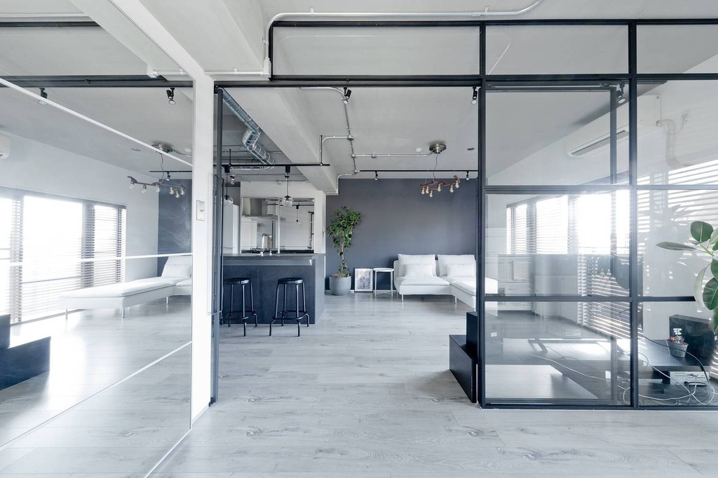 hacorenove style interior, industrial loft open-plan living space, black steel-framed glass partitions, exposed ceiling pipes and conduits, gray accent wall, light wood flooring, white sofa, minimalist monochrome palette, natural light