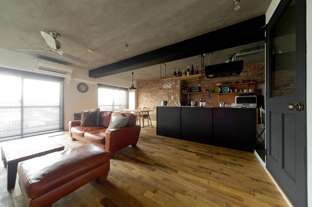 hacorenove style interior, industrial open-plan living kitchen, exposed brick wall, raw concrete ceiling, black steel beams, oak hardwood flooring, brown leather sectional sofa, open shelving, natural light, warm urban loft atmosphere