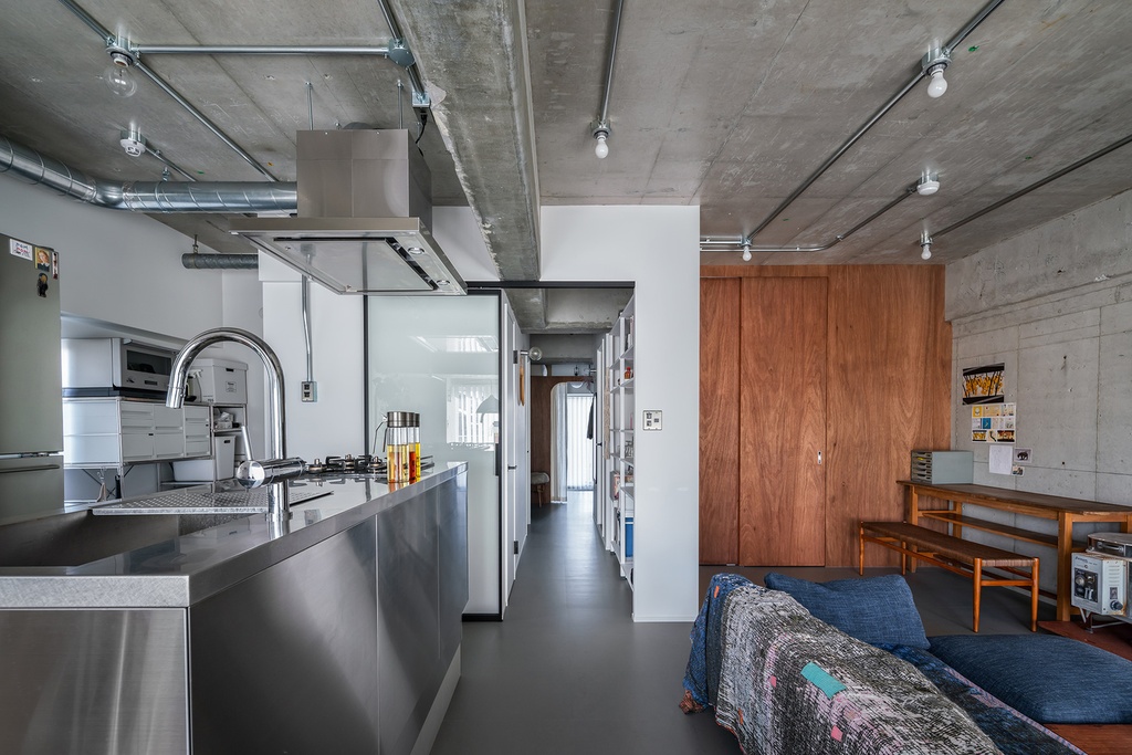 hacorenove style interior, industrial loft open-plan kitchen and living space, exposed concrete ceiling, stainless steel kitchen island, exposed conduit and pipes, wooden closet doors, gray floor, minimalist bare-bulb lighting