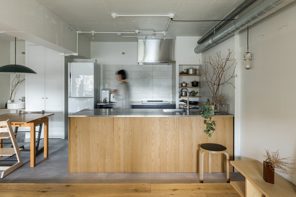 hacorenove style interior, Japanese industrial-minimalist open kitchen, oak island with stainless steel countertop, exposed ductwork, concrete ceiling, gray tile backsplash, natural light, warm neutral tones, dried branches