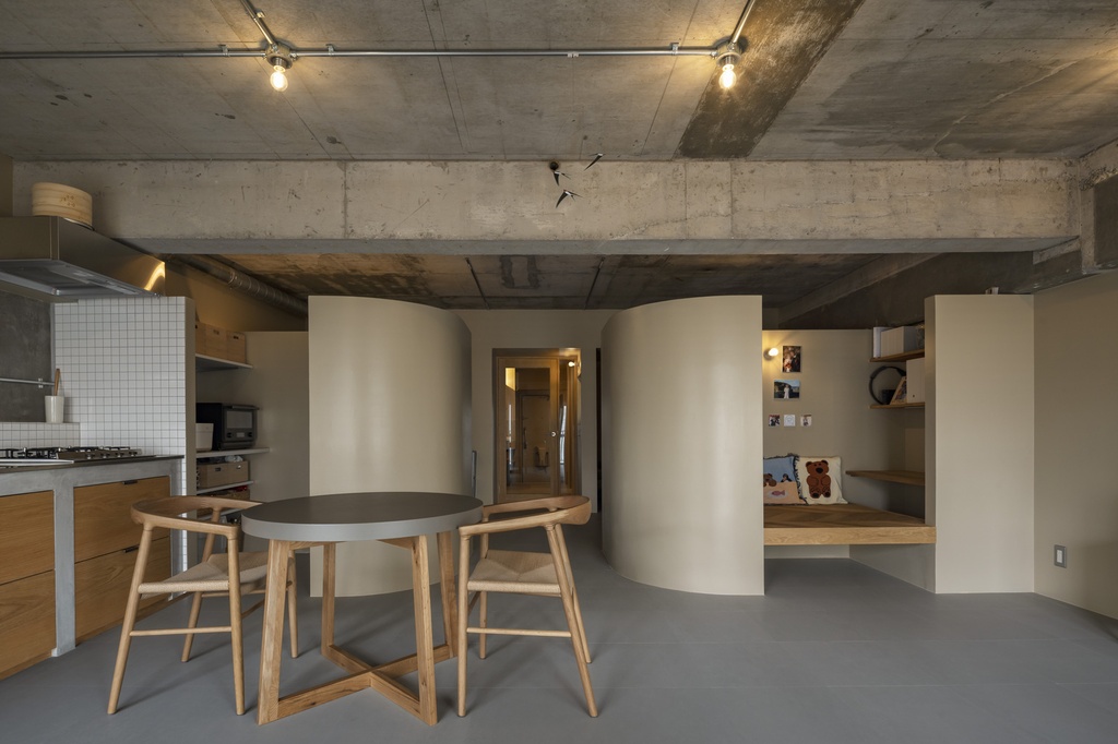 hacorenove style interior, open-plan apartment, exposed concrete ceiling, curved partition walls, gray floors, wooden dining set, track lighting, Japanese minimalist industrial, warm neutral tones, built-in reading nook