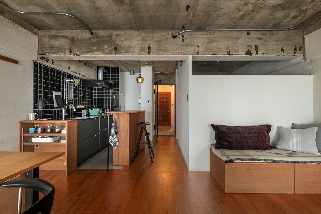 hacorenove style interior, industrial open-plan apartment, exposed concrete ceiling with conduit, black tile kitchen backsplash, warm wood flooring, built-in wooden daybed, minimal white walls, natural light