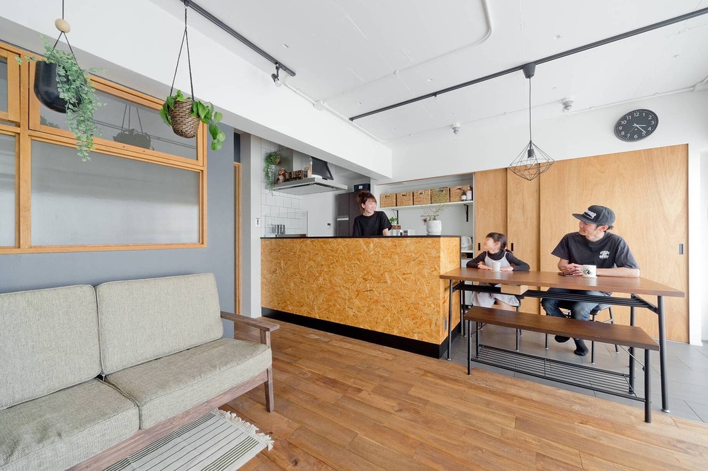 hacorenove style interior, open-plan living-dining-kitchen, OSB board kitchen island, blue-gray walls, wooden flooring, exposed ceiling pipes, black metal accents, hanging plants, natural light, Japanese industrial-casual atmosphere