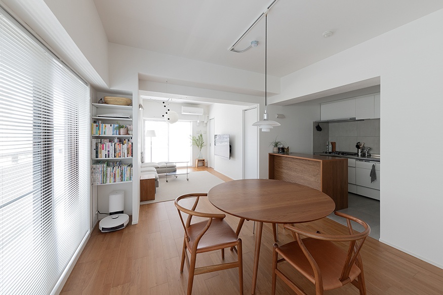 hacorenove style interior, minimalist Japanese apartment, open-plan dining and kitchen, warm oak flooring, round wooden table, wishbone chairs, white walls, pendant light, white blinds, soft natural light, clean Scandinavian-inspired aesthetic