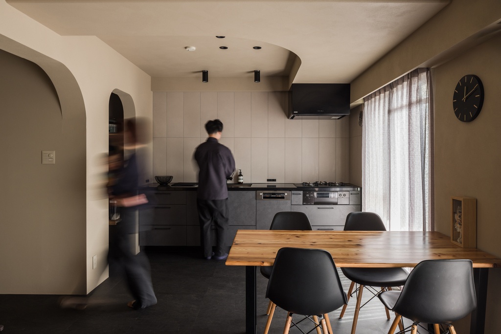 hacorenove style interior, modern Japanese kitchen-dining space, dark cement floors, taupe plaster walls, arched doorway, wooden dining table, black mid-century chairs, recessed spotlights, moody warm atmosphere, sheer curtains