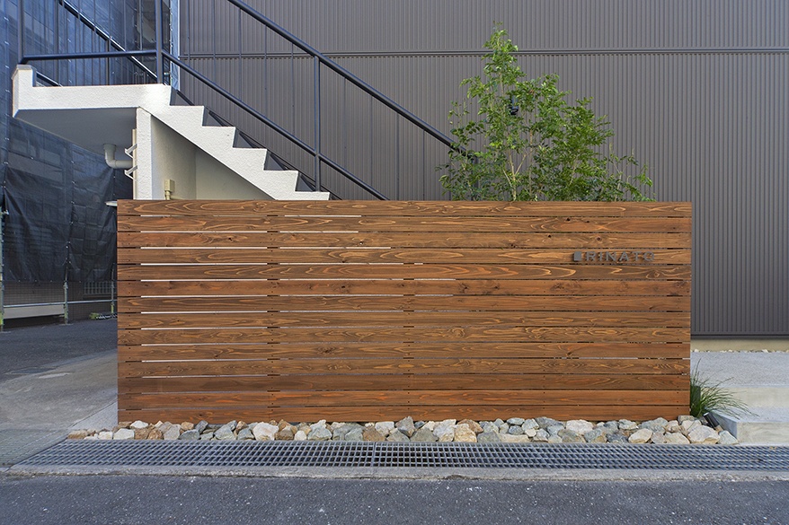 hacorenove style interior, exterior entrance facade, horizontal wood slat privacy screen, warm stained timber, river stones base, white concrete staircase, dark metal cladding, green accent tree, Japanese minimalist architecture, natural daylight