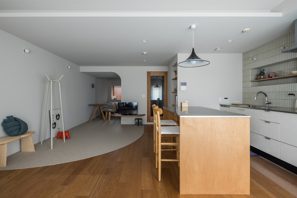 hacorenove style interior, open-plan kitchen and living room, curved archway, warm oak flooring, terrazzo accent floor, light wood island, sage green tile backsplash, black pendant light, minimalist Japanese aesthetic, soft neutral tones