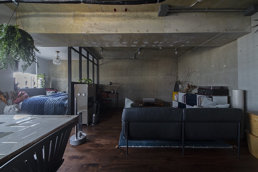 hacorenove style interior, industrial loft apartment, exposed concrete ceiling and walls, dark walnut hardwood floors, black steel-frame glass partition, moody dim lighting, hanging greenery, gray sofa, open-plan bedroom