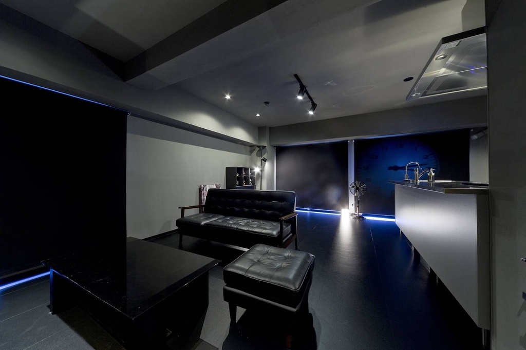 hacorenove style interior, dark moody open-plan living space, black leather sofa and ottoman, glossy black surfaces, blue LED accent lighting, grey walls, recessed spotlights, kitchen counter, ultra-modern masculine atmosphere