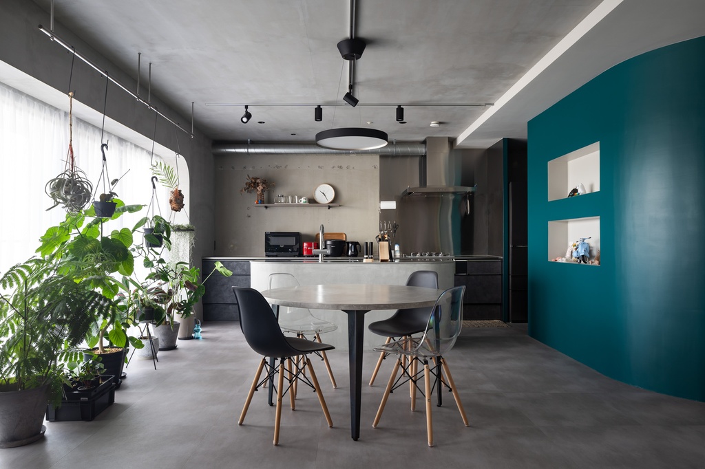 hacorenove style interior, industrial open-plan kitchen dining, raw concrete ceiling and floors, teal accent wall with recessed niches, hanging plants, black track lighting, mid-century chairs, moody urban atmosphere
