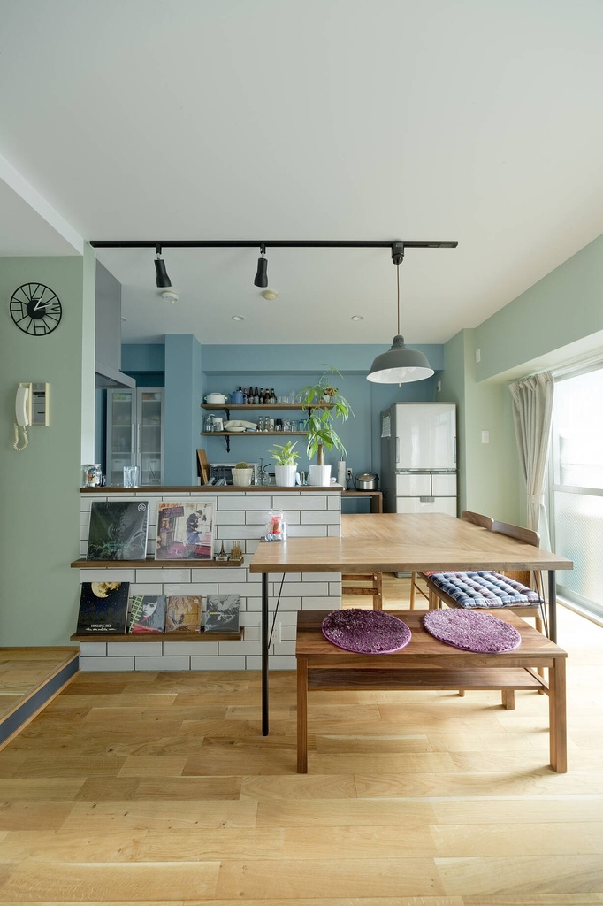 hacorenove style interior, open kitchen-dining room, sage green and blue accent walls, white subway tile counter, oak hardwood floors, walnut dining table with bench, black track lighting, Japanese casual living, natural daylight