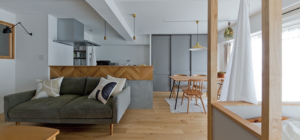 hacorenove style interior, open-plan living-dining-kitchen, light oak flooring, herringbone wood kitchen counter, gray-green sofa, white walls, exposed ceiling beams, brass pendant lights, gray panel doors, natural light, Japanese minimalist warmth