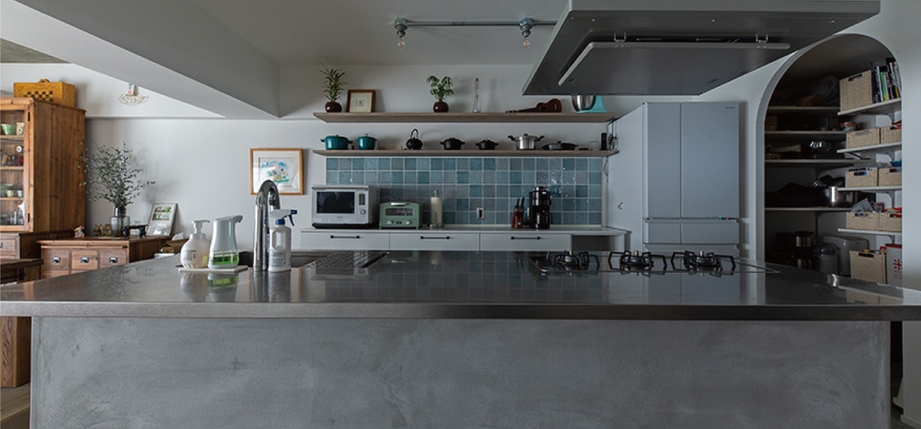 hacorenove style interior, Japanese kitchen with concrete island countertop, teal ceramic tile backsplash, open wooden shelving, arched pantry doorway, vintage wood furniture, white walls, natural daylight, lived-in warmth