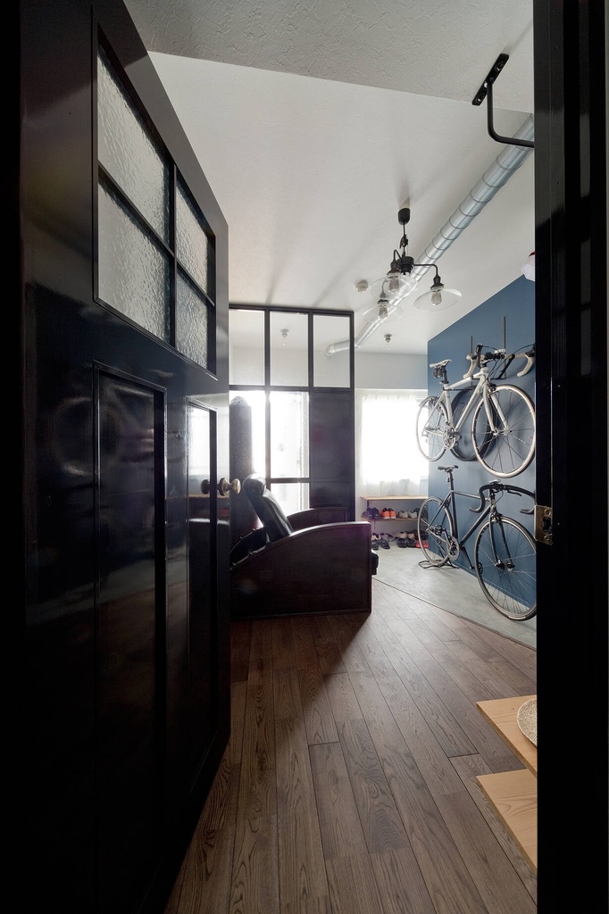 hacorenove style interior, industrial loft entryway, dark wood flooring, blue accent wall, wall-mounted bicycles, black steel-framed glass partitions, exposed ductwork, leather armchair, natural light, urban masculine atmosphere