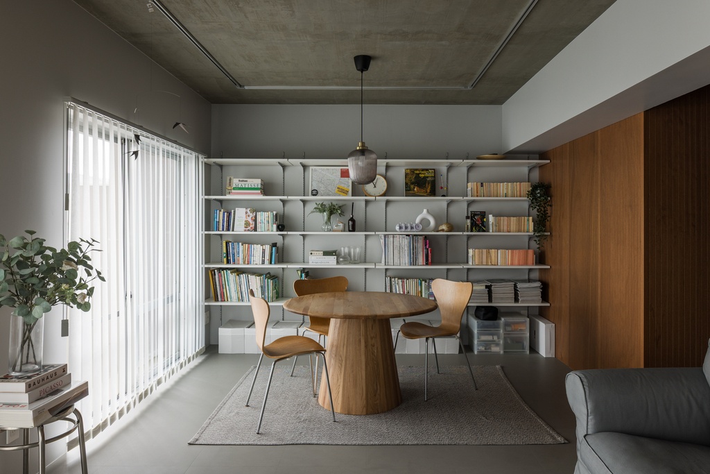 hacorenove style interior, modern dining area, exposed concrete ceiling, gray walls, wall-mounted metal shelving with books, round oak pedestal table, bentwood chairs, warm wood accent wall, soft natural light, minimalist industrial atmosphere