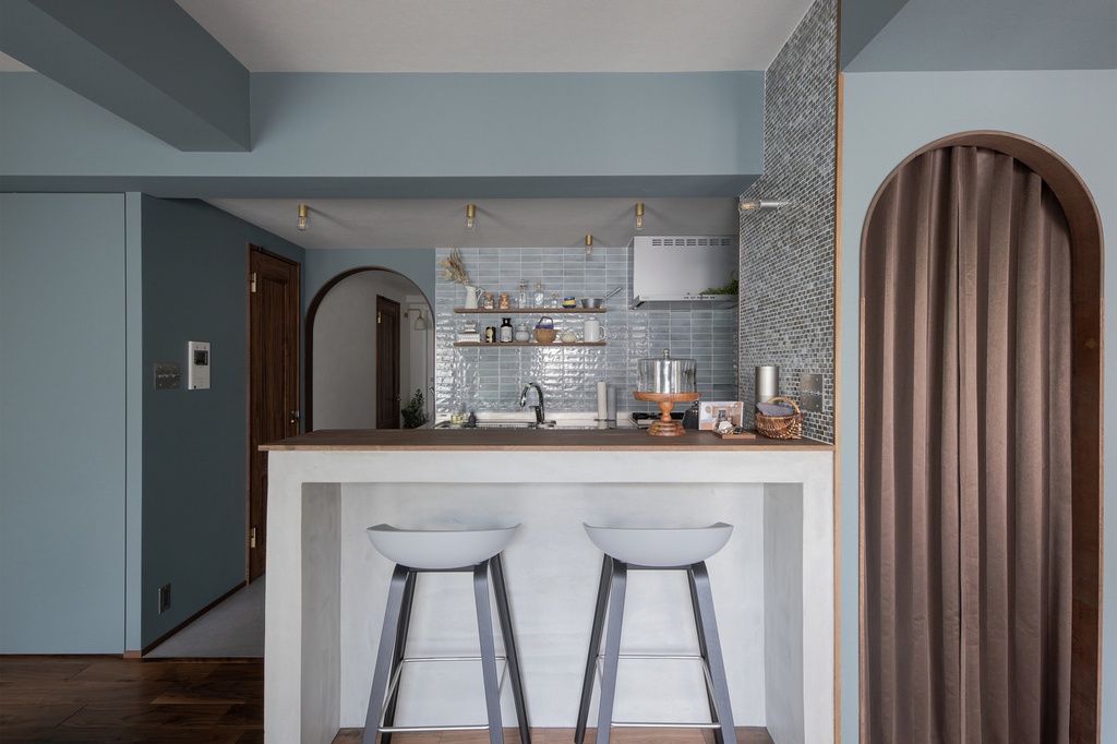 hacorenove style interior, open kitchen with breakfast bar, dusty blue walls, glossy blue subway tile backsplash, arched doorways with curtains, white concrete counter, dark wood floors, brass track lighting, warm cozy atmosphere