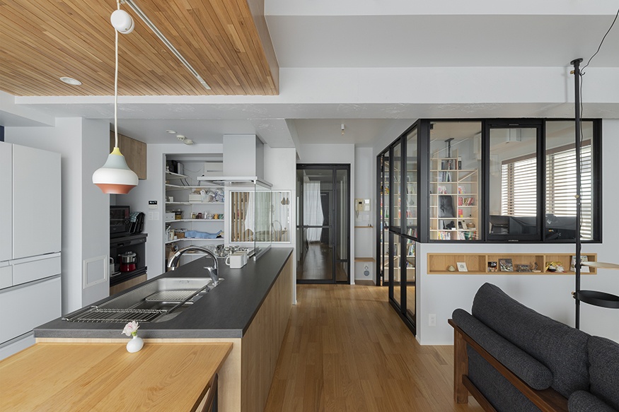 hacorenove style interior, open-plan kitchen-living space, warm oak flooring, wood-paneled ceiling accent, black countertops, black steel-framed glass partitions, white walls, colorful pendant lights, Japanese modern minimalist atmosphere, natural daylight