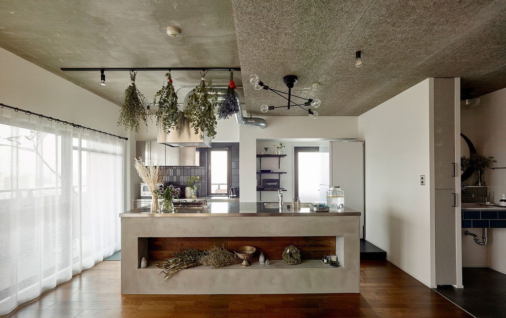 hacorenove style interior, open-plan kitchen with concrete ceiling and island counter, warm wood flooring, hanging dried herbs, industrial chandelier, white walls, blue tile accents, natural light, sheer curtains, botanical rustic atmosphere