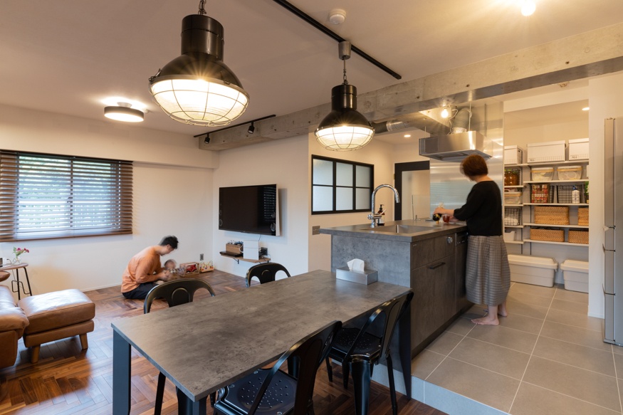 hacorenove style interior, open-plan kitchen-dining-living room, concrete countertops, exposed concrete beams, industrial black pendant lights, dark wood herringbone flooring, gray tile kitchen floor, white walls, black metal chairs, warm natural lighting