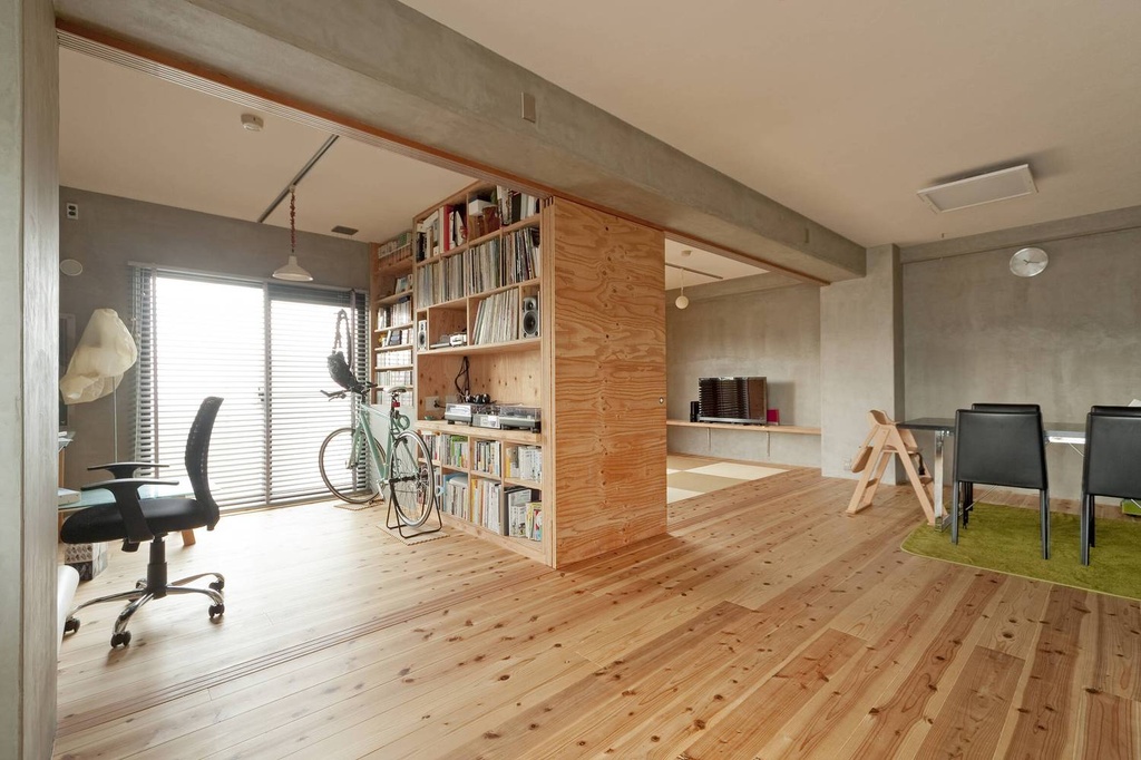 hacorenove style interior, open-plan Japanese apartment, exposed concrete beams and walls, natural pine wood flooring, plywood bookshelf room divider, minimalist industrial aesthetic, natural daylight, warm neutral tones
